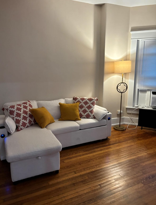 A living room featuring a white sectional sofa, with a mix of patterned and solid cushions, a wooden floor, a tall lamp, blinds, and a small cabinet.