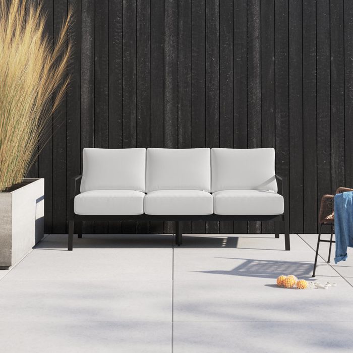 A modern outdoor patio featuring a sleek three-seat sofa set against a dark wood panel wall, complemented by a minimalist design and potted plants nearby.