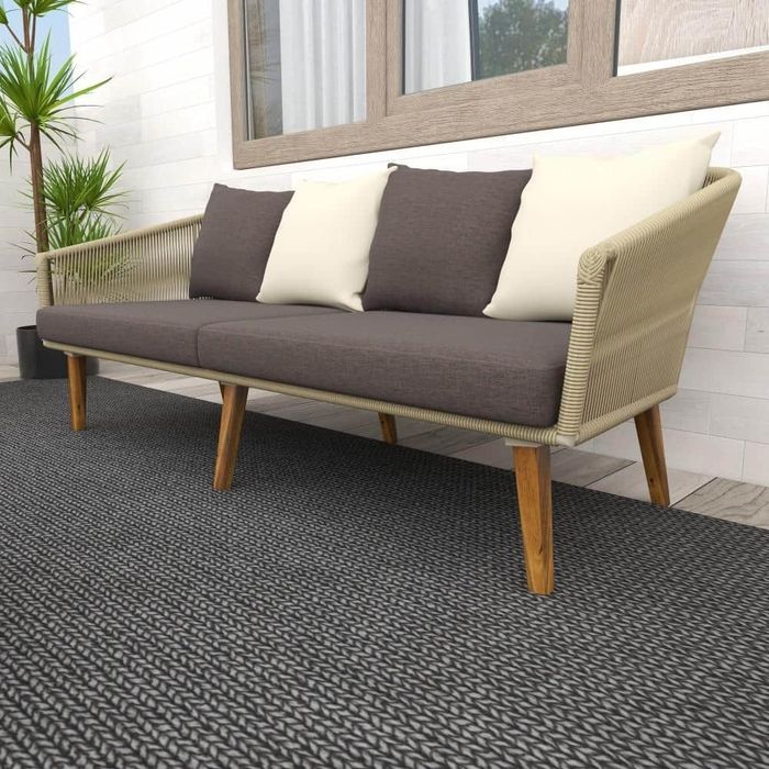 A contemporary patio sofa with wooden legs and beige frame, paired with beige and brown cushions. It's placed on a textured rug, near a white wall adorned with windows and indoor plants.