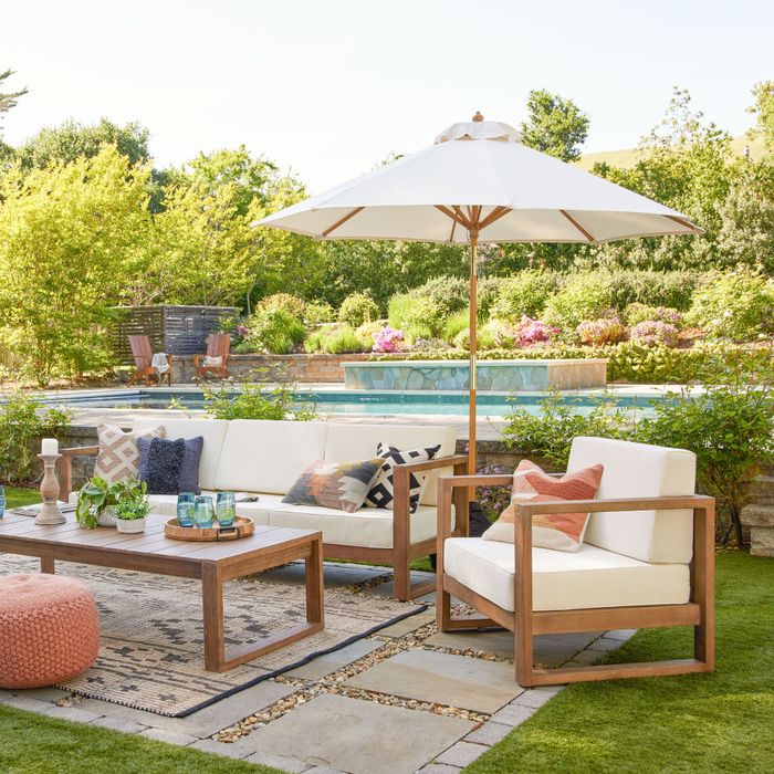 An outdoor patio setup with a wooden lounge set, white cushions, a coffee table, a pouf, and a large umbrella, all beside a poolside garden.