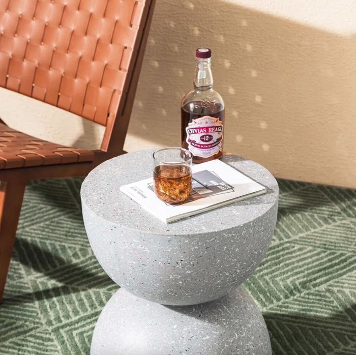 A grey hourglass-shaped stool sitting next to an outdoor chair, with a drink and a book placed on top.