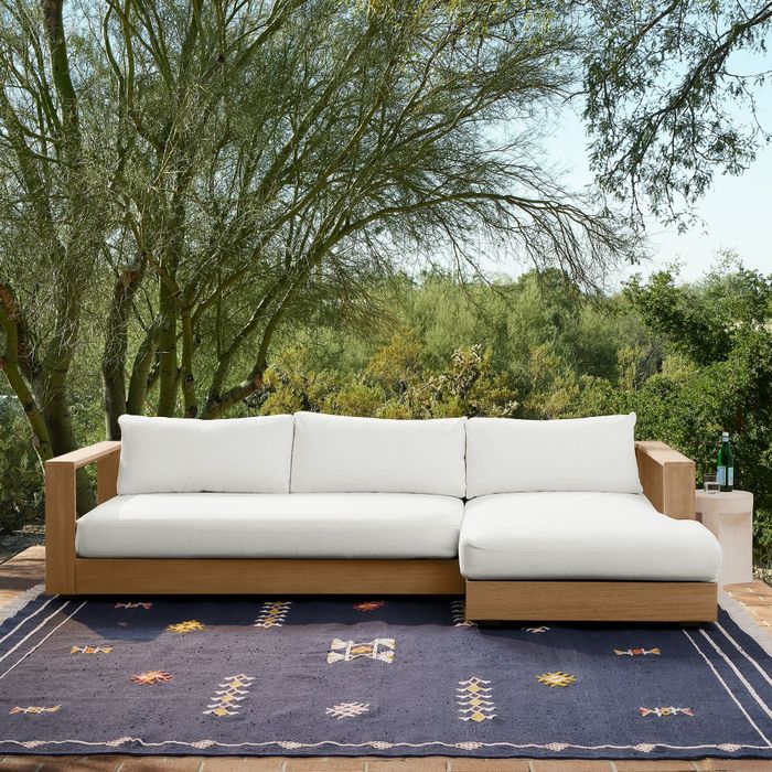 A wooden outdoor sectional sofa with white cushions, placed on a decorative rug, surrounded by trees.