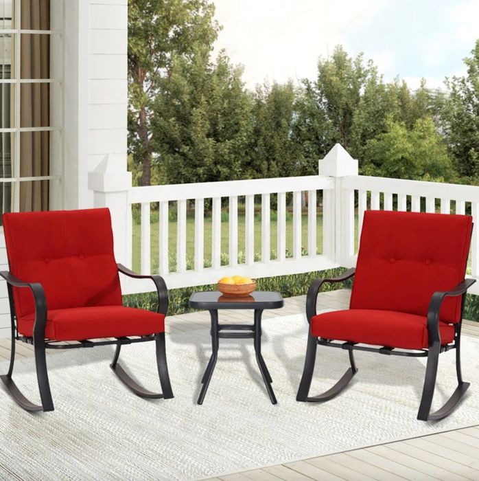 A set of black metal rocking chairs with red cushions and a small table. The set is placed on a white porch with a scenic view of the yard and a wooded area.