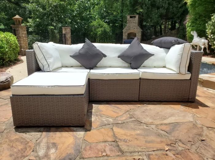 A dark wicker sofa with light cushions, placed on a stone patio with trees gently swaying in the background.