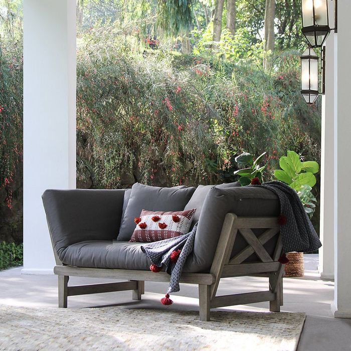 A modern dark cushioned loveseat on an outdoor patio, complete with patterned pillows and a cozy knit throw, all set against a lush garden backdrop.