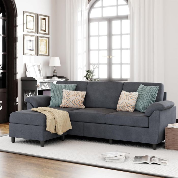 A modern living room features a dark sectional sofa adorned with patterned and textured pillows, placed near a large window with sheer curtains and framed art on the walls.