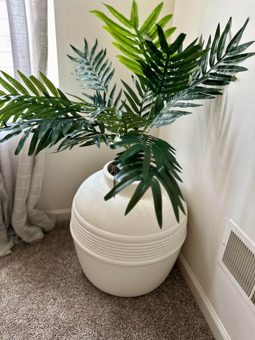 A large white ceramic pot in a room corner, filled with lush artificial palm leaves, placed on a carpet near a window with drawn curtains.