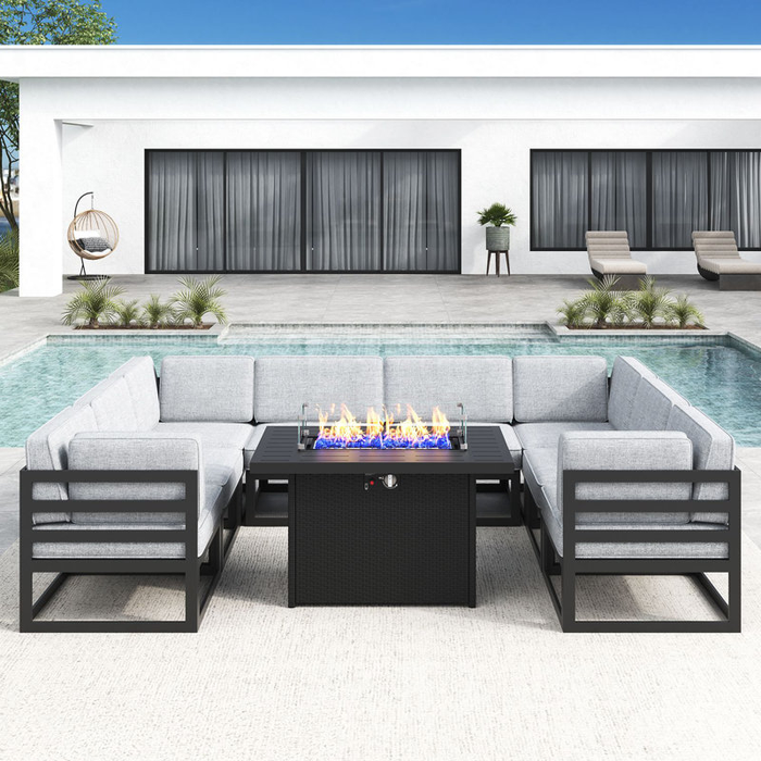 An outdoor patio set with gray cushioned seating around a rectangular fire pit, placed near a modern house with a pool and lounge chairs.