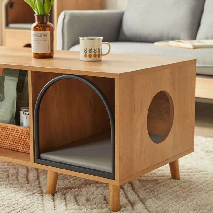 A wooden coffee table with a built-in pet bed, featuring a round hole on the side. On top, there’s a cup and a bottle, with a sofa in the background.