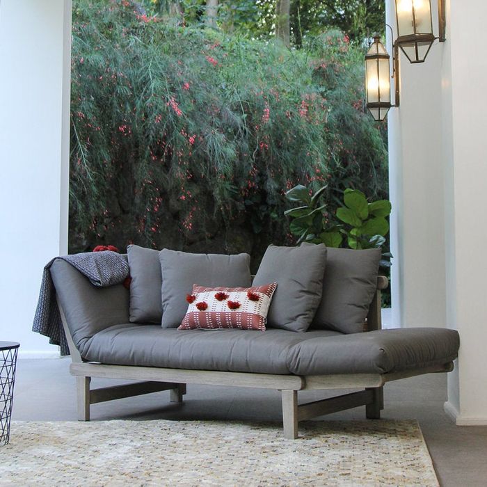 A contemporary gray daybed with soft cushions and a red-patterned pillow, positioned in a patio area surrounded by greenery and stylish lanterns.