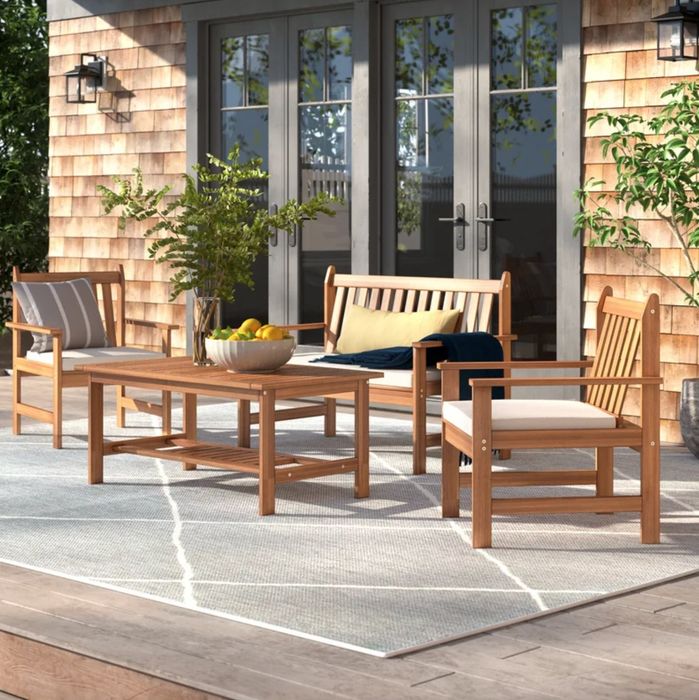 A light wood coffee table, accompanied by chairs and a loveseat with soft, light-colored cushions. The entire set rests on a light outdoor rug, set up on a deck.