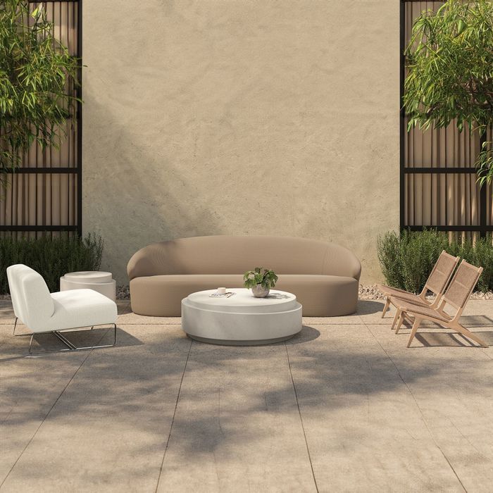 A modern outdoor patio setup with beige sofas, a white armchair, a round table, two wooden chairs, and plenty of plants set on a tiled floor.