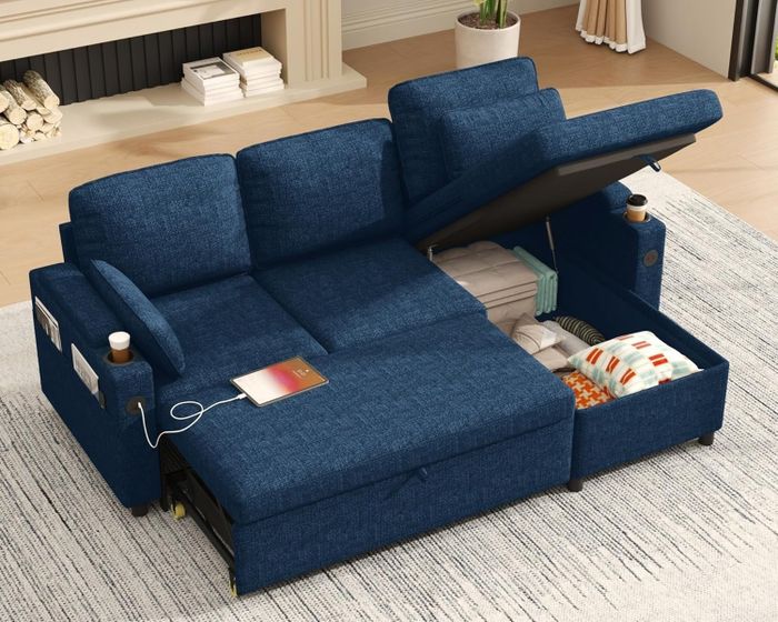 A convertible blue sectional sofa with hidden storage, revealing blankets and a book, placed on a living room rug.