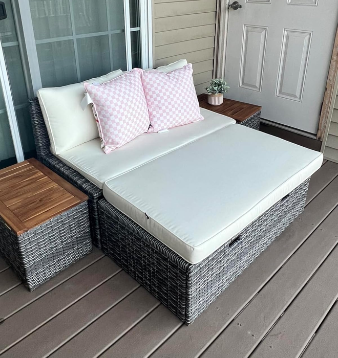 A stylish outdoor wicker daybed adorned with white cushions and pink checkered pillows, set on a wooden deck beside small side tables.
