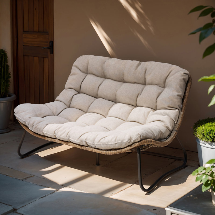 A cushioned outdoor loveseat with a wicker frame and metal legs, placed on a patio surrounded by plants, with a wooden door in the background.