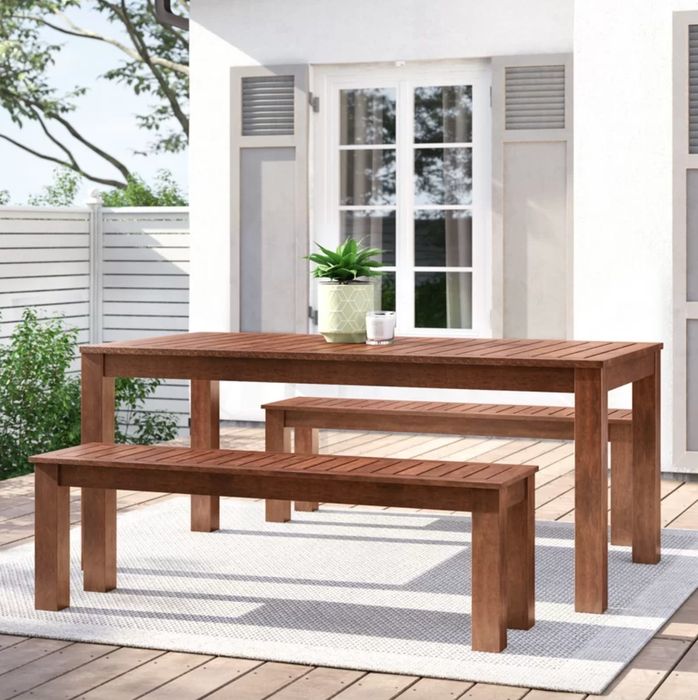The medium wood table and benches are placed on a beige rug in a stylishly decorated porch area, creating a cozy and inviting outdoor dining space.