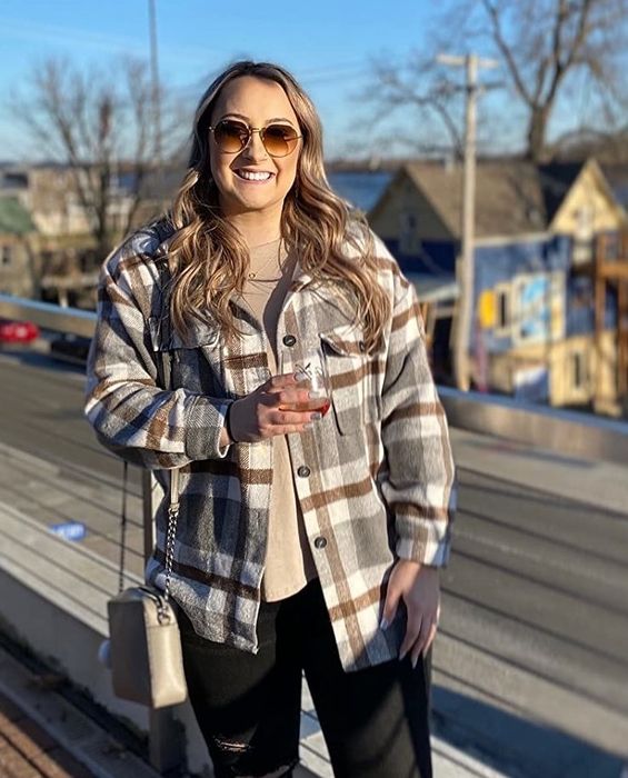 A woman enjoys the outdoors, wearing a casual flannel jacket and sunglasses, holding a drink while standing on a balcony overlooking a street.