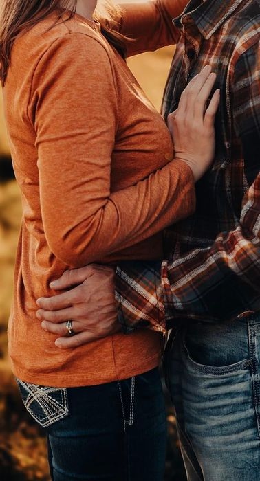Two people dressed casually share an embrace; one wears an orange shirt, while the other has on a plaid shirt and jeans. Their faces are hidden from view.