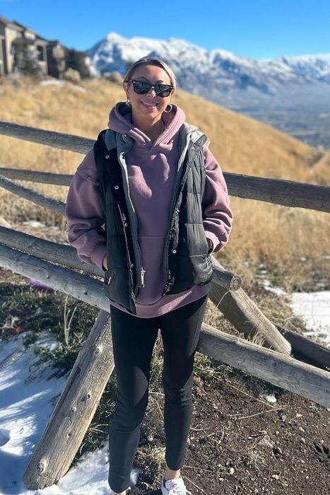 A reviewer stands on a snowy mountain slope near a wooden fence, wearing sunglasses, a pink hoodie, and a black vest, with a breathtaking mountain view in the background.