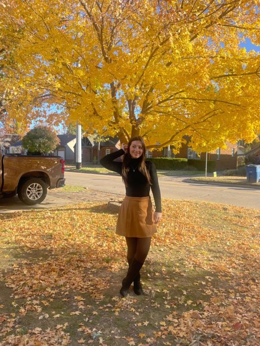 A person smiles beneath a tree with vibrant autumn leaves, dressed in a black long-sleeve bodysuit and a tan skirt, standing in a residential neighborhood.