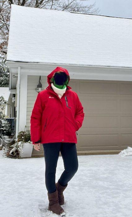 A person stands in a snowy driveway, bundled up in a red coat, black leggings, brown boots, and a green scarf, with a cozy house in the background.