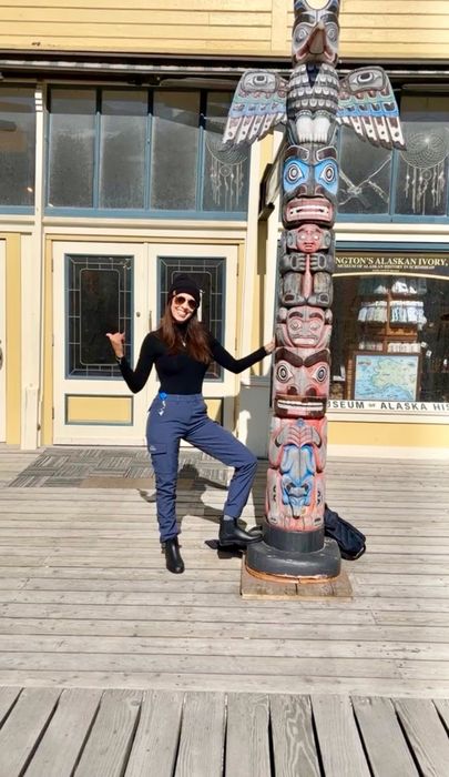 A reviewer strikes a fun pose next to a totem pole on a wooden deck, sporting a black top and blue pants.