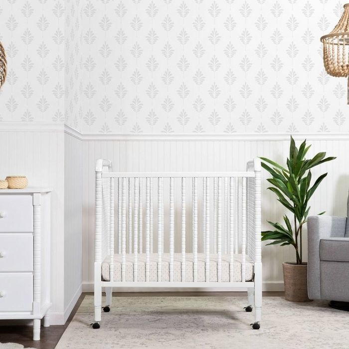 A white mini crib set up in a nursery with stylish geometric wallpaper in the background.