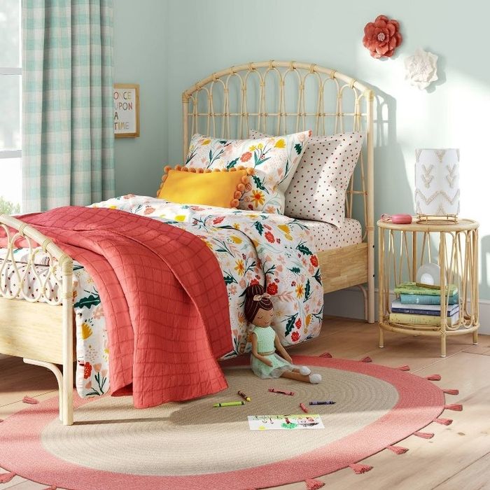 A child’s bedroom with a neatly made bed, an oval rug, a doll, and a rattan bedside table, exuding a calm and organized vibe.