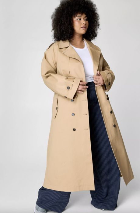 A person with curly hair dons a beige trench coat, white shirt, and wide-leg striped pants.