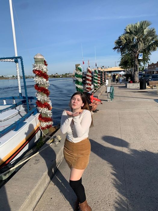 A person stands on a dock next to a decorated boat, smiling with hands beneath their chin, wearing a white top and tan skirt.