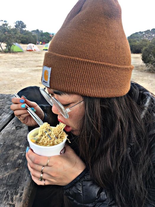 A reviewer, wearing a beanie and glasses, enjoys a meal of instant noodles outdoors, eating with a fork and maintaining a focused expression.