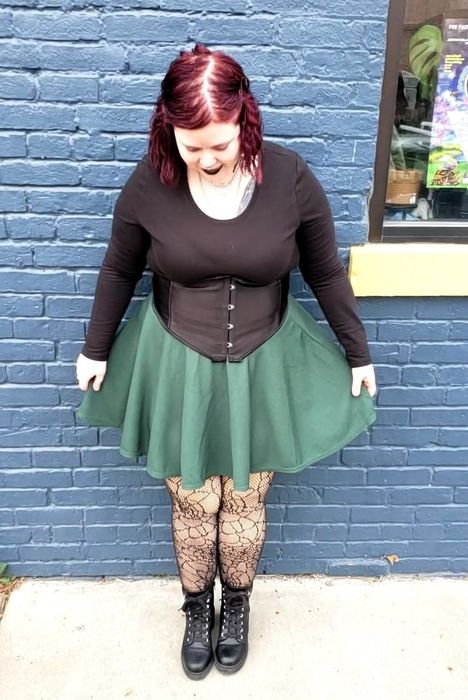 A reviewer stands against a brick wall, wearing a green skirt, black top, lace stockings, and boots, looking down.