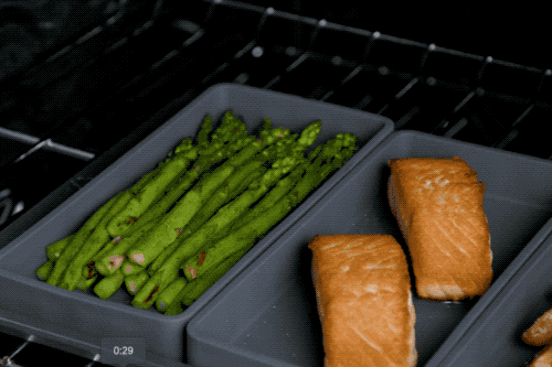A model removing asparagus from the oven and placing salmon and potatoes back inside to continue cooking.