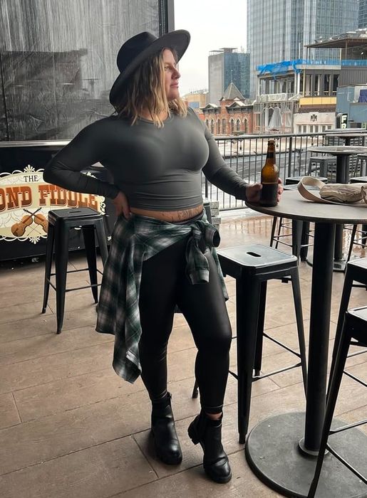 A person dressed casually, with a hat, a gray long-sleeve crop top, and a plaid shirt tied around their waist, stands on a bar patio holding a drink, with a cityscape in the background.