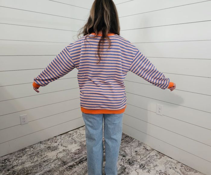 A person dressed in a striped sweater and jeans, standing with arms outstretched, facing away in a room with white-paneled walls.