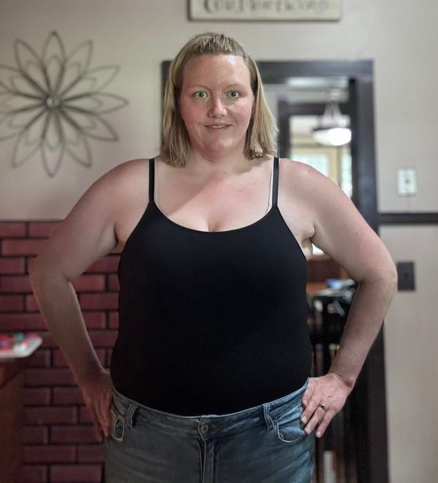A person stands indoors, confidently smiling at the camera, dressed in a black top and grey pants, hands on their hips.