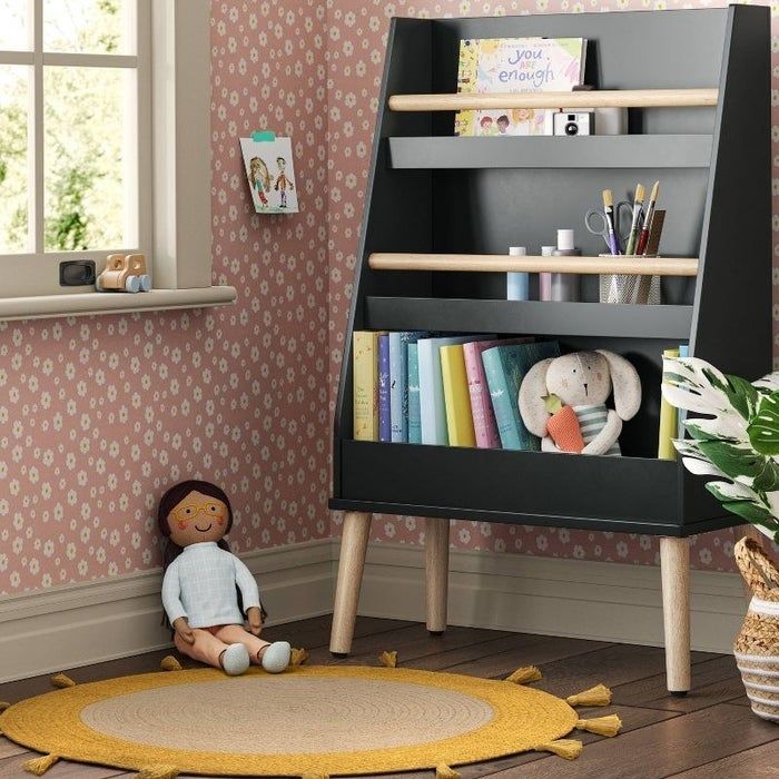 A sleek charcoal grey three-shelf bookshelf packed with books in a kid's bedroom, accompanied by a sun-shaped rug and a cuddly plush doll.