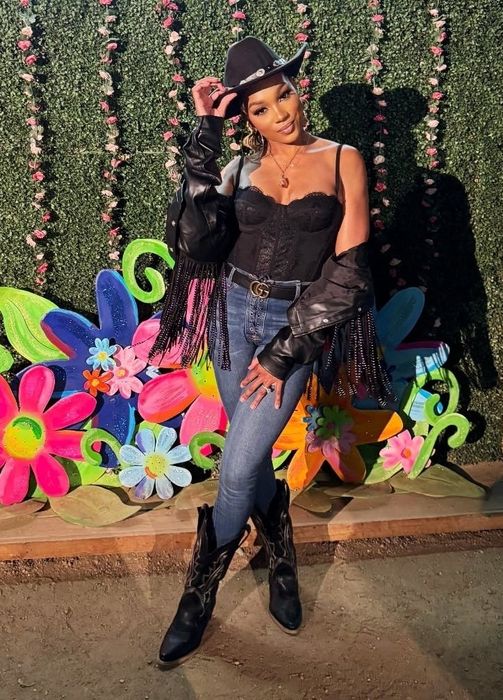 A person strikes a pose in a cowboy hat, black lace top, jeans, and boots in front of a #RodeoUncorked event backdrop adorned with colorful flowers.