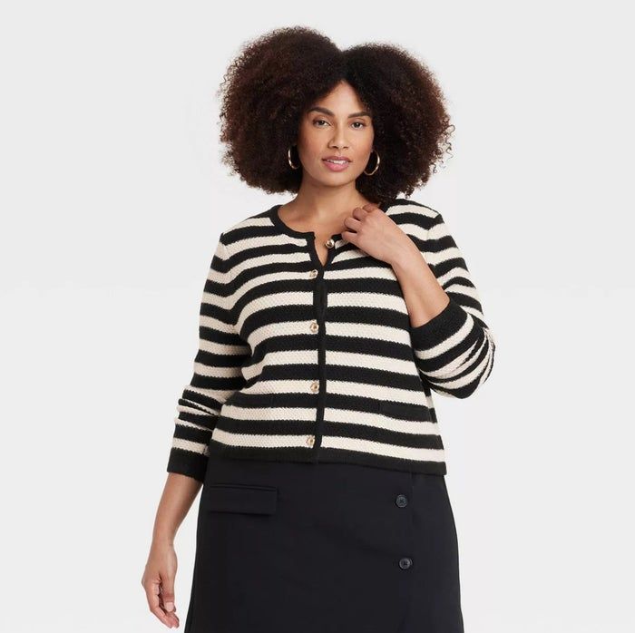 A model with curly hair wearing a black and white striped cardigan and a black skirt, striking a pose against a plain backdrop.