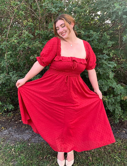 A woman smiles while posing outdoors in a flowing, off-shoulder red dress, holding the sides of her skirt