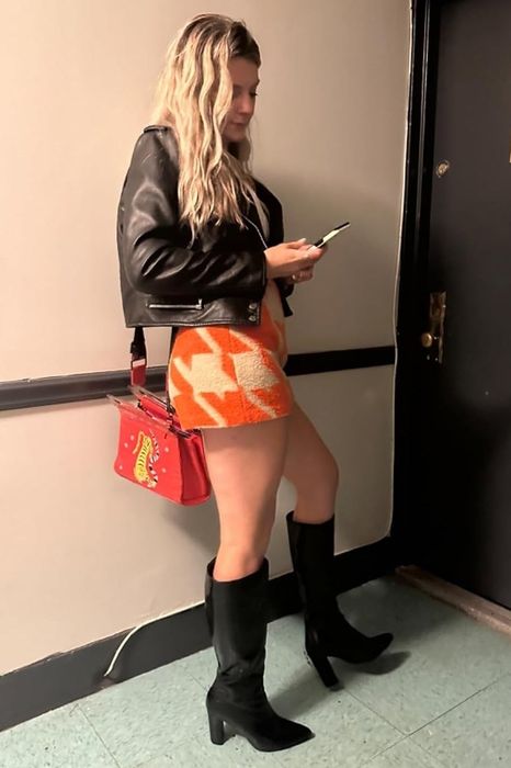 Reviewer standing by a door, wearing a leather jacket, a patterned skirt, black boots, and holding a red bag, while looking at their phone