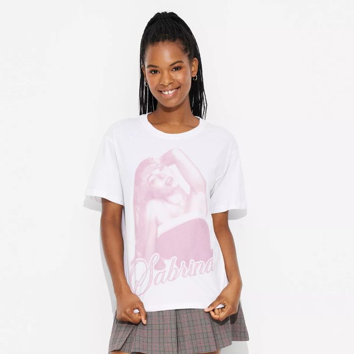 A smiling woman wearing a white T-shirt featuring a large graphic of Sabrina, paired with a plaid skirt, standing against a simple background.