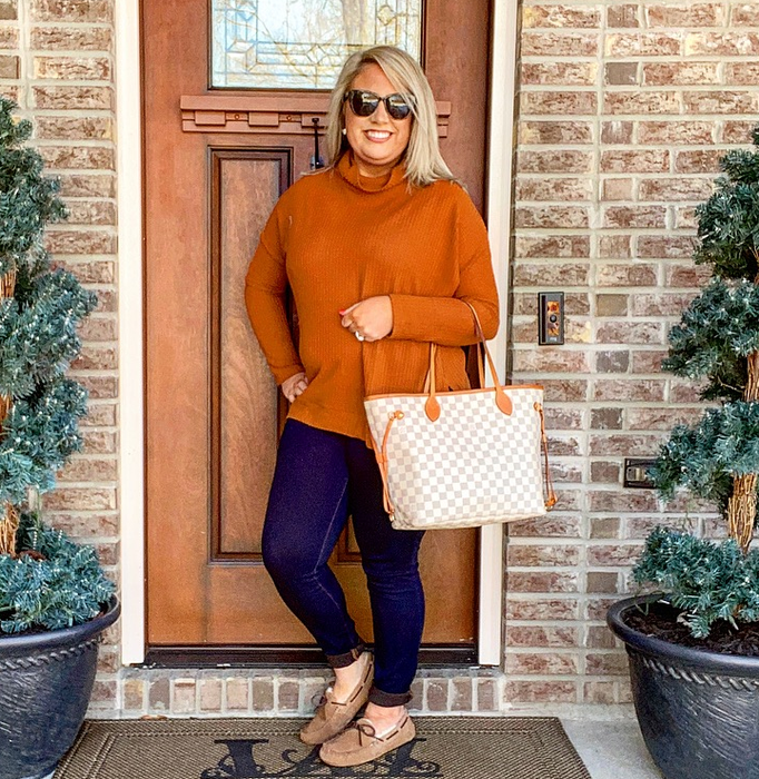 A reviewer strikes a pose in front of a door flanked by evergreen plants, wearing a caramel tunic sweater, jeans, ankle boots, sunglasses, and carrying a checkered tote bag.