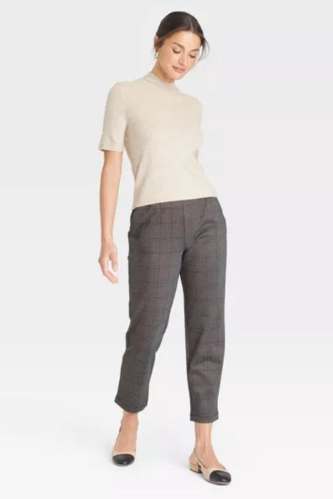 A person in a short-sleeve top and checkered pants, standing against a plain backdrop with a relaxed, slightly leaned posture.