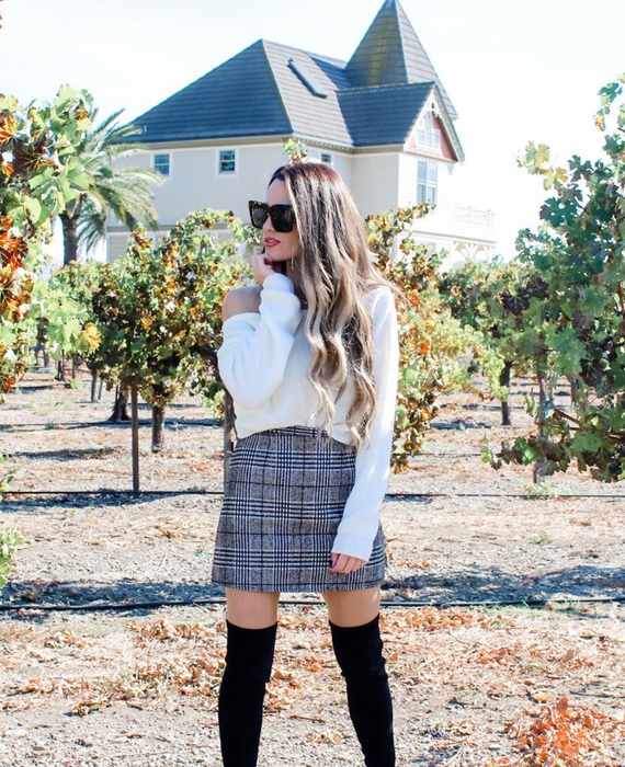 A reviewer poses in a vineyard, wearing a white off-shoulder sweater paired with a plaid mini skirt and black thigh-high boots.
