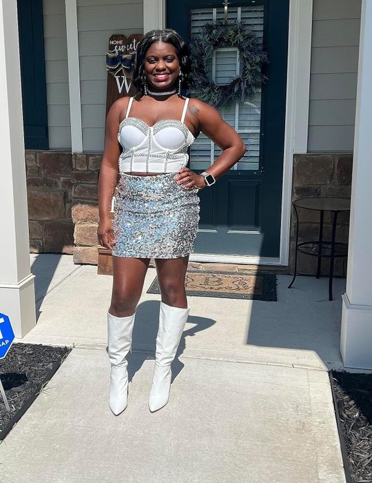 A person standing outside a house wearing a pearl-studded corset top, a shimmering sequin skirt, and white knee-high boots