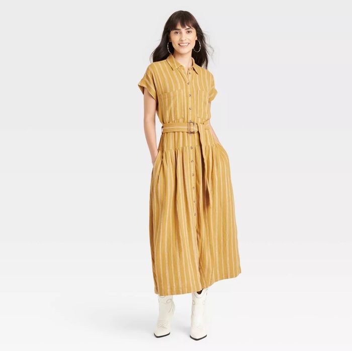 A person stands smiling, wearing a belted, ankle-length striped button-down dress with short sleeves, paired with boots