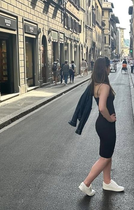 A woman stands confidently on a narrow city street, dressed in a black sleeveless dress, paired with white sneakers, while casually holding a jacket over her shoulder.