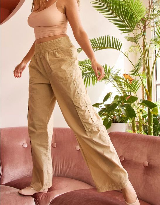 A model strikes a pose wearing beige cargo pants and a light tank top, sitting on a pink velvet sofa surrounded by lush greenery.