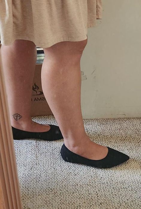 Person modeling black pointed flats on a carpet, displaying a diamond leg tattoo on the left leg